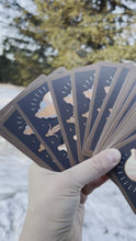 Load and play video in Gallery viewer, The backside of the Gentle Tarot Linen Edition cards, fanned out, held in one hand, with snow and trees in the background.
